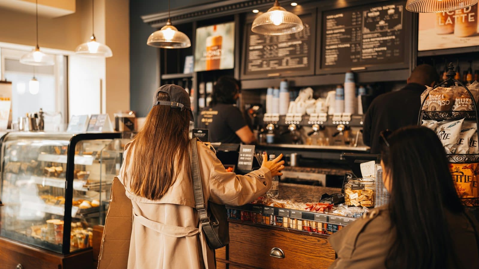 A customer at a café counter reaching toward the till, with the pastry case, the menu boards and the espresso machine behind her, and another customer waiting.