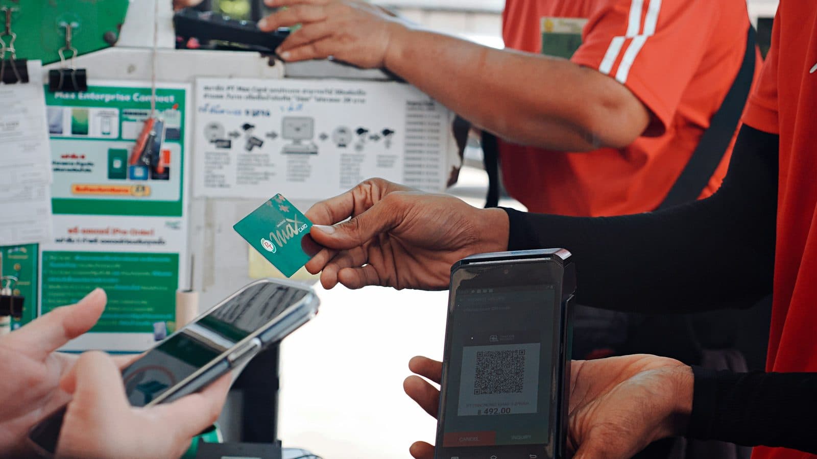 At a small kiosk counter, one hand holds out a green plastic loyalty card while another holds a card terminal showing a QR code, with a customer's phone in the foreground.