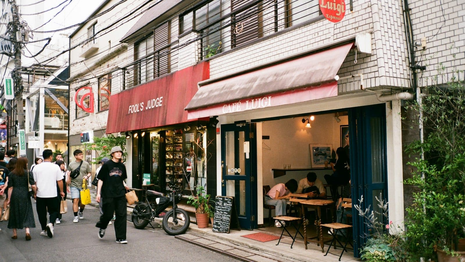 Two small shopfronts side by side on a narrow street, a clothing store under a red awning and Cafe Luigi next door with customers at a table just inside the open doorway, as pedestrians walk past.