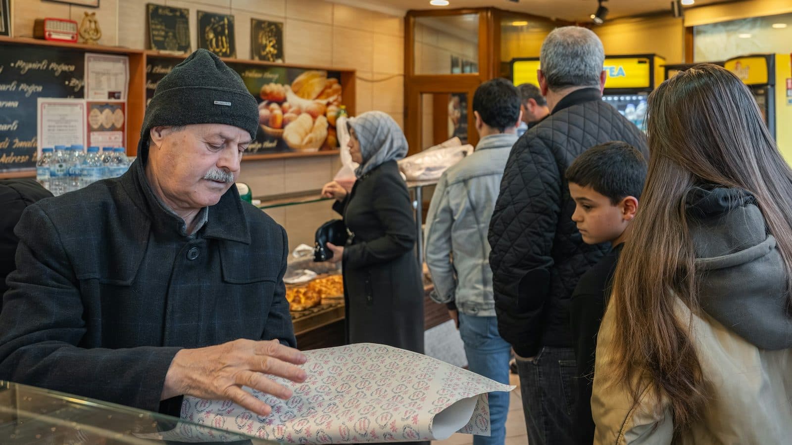 An elderly baker smoothing a sheet of wrapping paper over the display case of a small bakery while a queue of customers waits to be served.