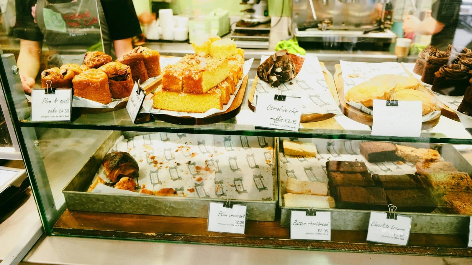 A glass cafe display case of cakes, brownies, shortbread and a lone croissant, each tray labelled with a handwritten price card, with a member of staff working behind it.