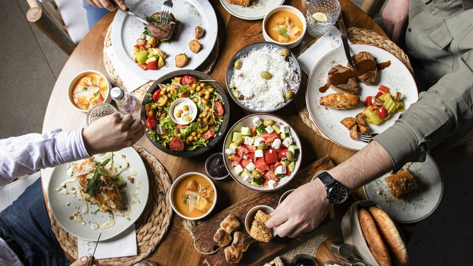 An overhead view of a round wooden restaurant table crowded with shared plates of salad, soup, bread and grilled meat, with three diners reaching in from the edges of the frame.
