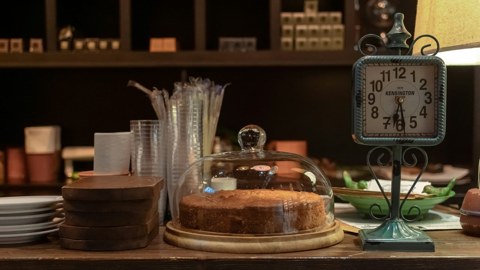 A dark cafe counter holding a sponge cake under a glass dome, a stack of white plates and wooden boards beside it, and a standing clock face reading half past six.
