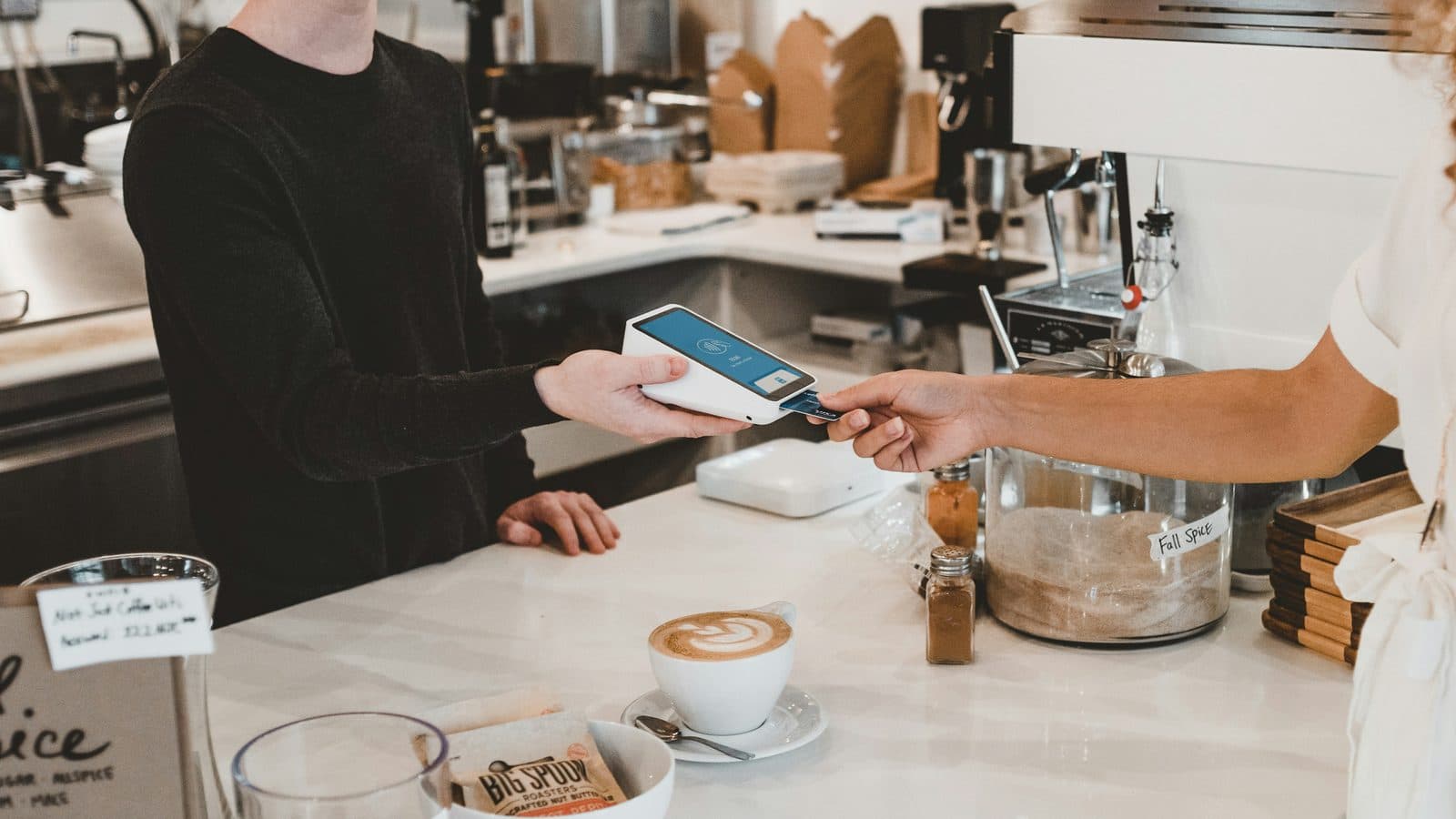 A barista holds out a small white card terminal across a marble cafe counter while a customer taps a card against it, with a finished latte and jars of spice sitting between them.