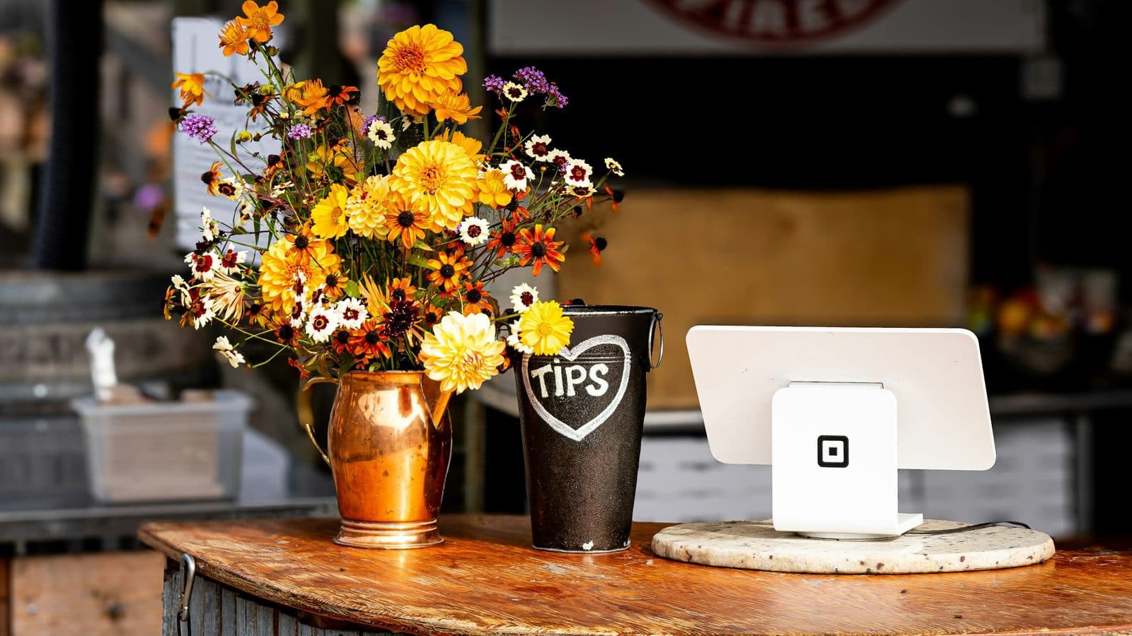 A wooden checkout counter at an outdoor pizza shop, holding a copper jug of orange and yellow flowers, a chalk-marked tips tin, and a single card reader on a marble slab.
