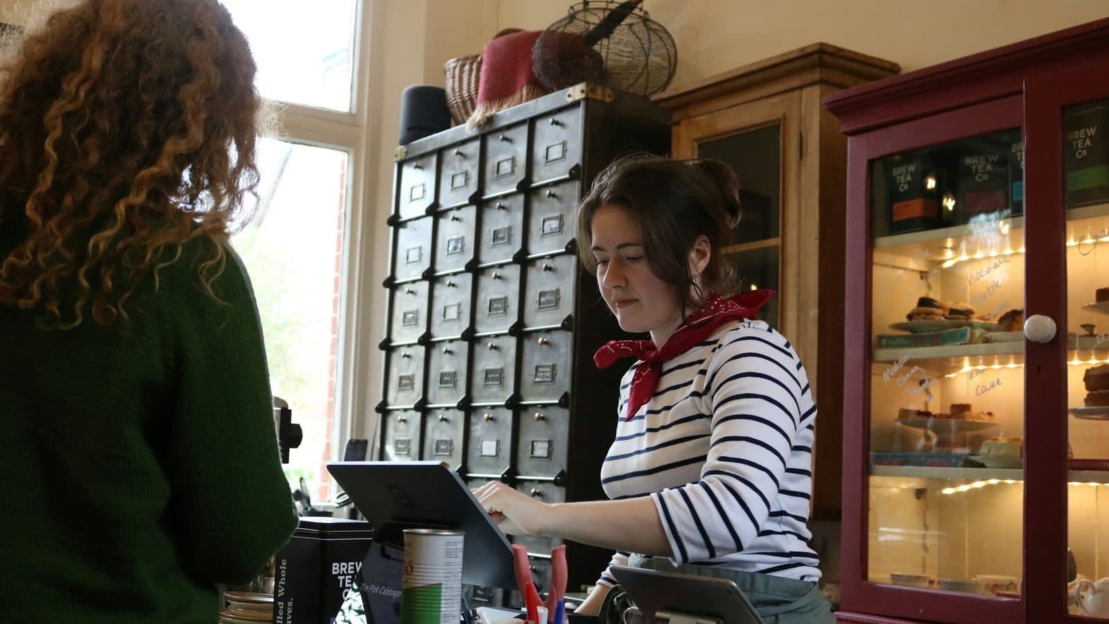 A woman in a striped top and red neckerchief works a tablet till behind the counter of a small tea shop while a customer waits, with cake stands lit in a cabinet beside her.