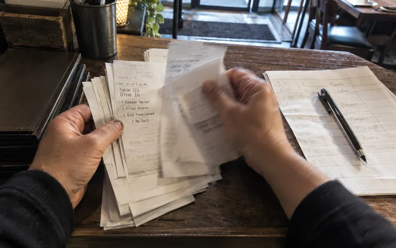 Hands flipping through printed order tickets at a restaurant host stand