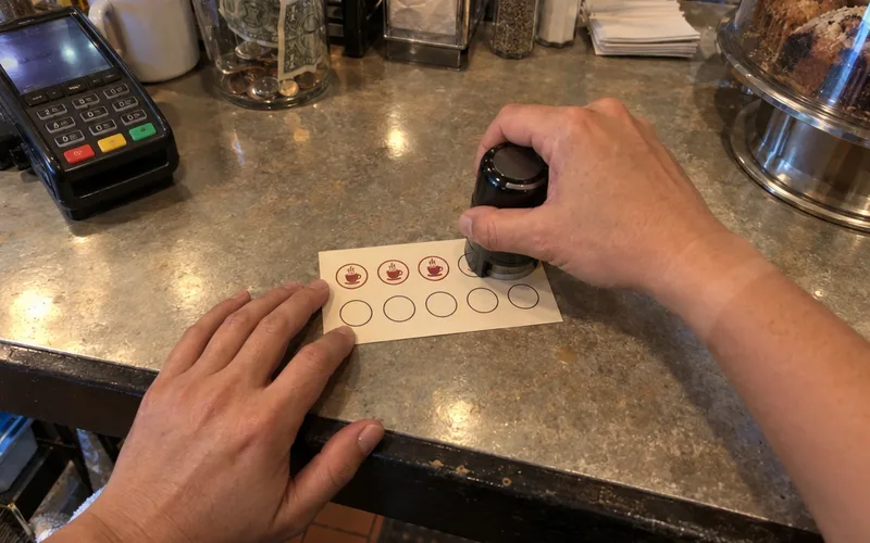 A cashier's hands pressing a stamp onto the fourth circle of a paper loyalty punch card at a diner counter